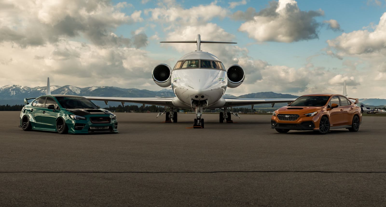 Austin's green STI and Colby's orange WRX parked either side of a private jet on the tarmac, mountains behind